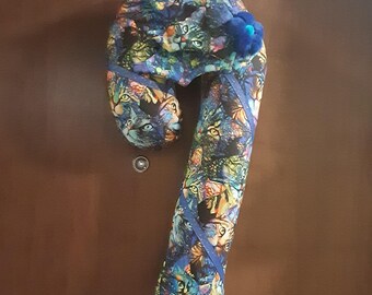 May include: A candy cane shaped fabric door hanger featuring a colorful print of cat faces in shades of blue, green, and orange. The soft, stuffed decoration includes a dark blue fabric ribbon accent and a small blue fabric flower at the top.