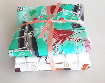 May include: A stack of folded fabric pieces tied with a peach-colored ribbon. The top fabric is teal with a pattern of red cocktails, candy canes, and white snowflakes. The bottom fabric is white with a print of various beverage glasses and text.