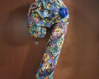 May include: A candy cane shaped fabric draft stopper featuring a colorful, multi-tonal cat print pattern in shades of blue, green, and orange. The item is accented with a small, dark blue fabric flower and thin purple ribbon trim along the length.