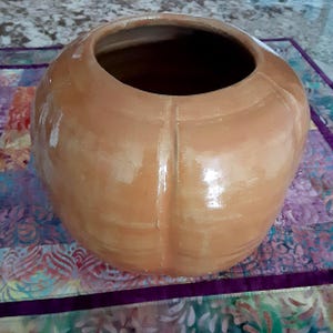 May include: A round, glazed ceramic vessel with a light brown, terracotta-toned finish. The exterior features vertical indentations that create a segmented, pumpkin-like appearance. It has a wide, circular opening at the top and a smooth, reflective surface texture throughout the body.
