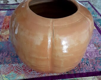 May include: A round, glazed ceramic vessel with a light brown, terracotta-toned finish. The exterior features vertical indentations that create a segmented, pumpkin-like appearance. It has a wide, circular opening at the top and a smooth, reflective surface texture throughout the body.
