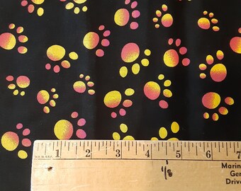 May include: A black fabric featuring a repeating pattern of paw prints in gradient shades of yellow and pink. A wooden ruler is positioned across the bottom, showing the paw prints measure approximately 0.5 to 0.75 inches in width.