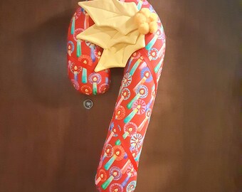 May include: A plush candy cane decoration featuring a red fabric base with a repeating pattern of colorful candles and circular designs. The top of the candy cane is adorned with three large, pale yellow fabric leaves and small yellow pom-poms.