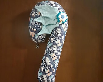 May include: A blue and white candy cane shaped pillow with a blue bow and a white and blue pom-pom on top. The pillow has a pattern of palm trees and beach houses.
