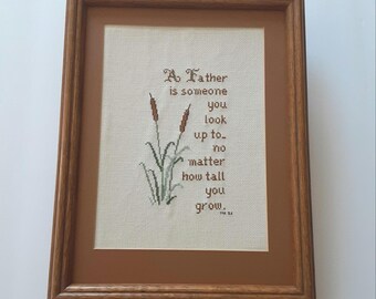 May include: Framed cross-stitch artwork with a brown wooden frame. The artwork features the text "A Father is someone you look up to..." with stitched cattails and greenery. The frame is rectangular.