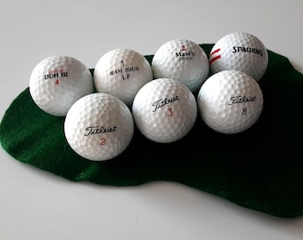 May include: Seven white golf balls with dimpled surfaces, featuring various brand names and numbers including Dunlop DDH III 4, Ram Tour LP 1, Maxfli DDH II 2, Spalding, and Titleist 2, 3, and 8, arranged on a dark green felt-like fabric piece.