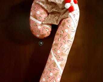 May include: A candy cane shaped fabric pillow featuring a pink background with a repeating pattern of gingerbread men, candy canes, and holly leaves. A white ribbon wraps around the pillow, accented by a brown fabric bow and red and white pom-poms.