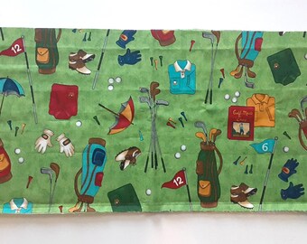 May include: A rectangular piece of green fabric featuring a repeating pattern of golf-themed illustrations including golf bags, clubs, balls, tees, umbrellas, gloves, shoes, shirts, and a book titled Golf More Often with a red flag numbered 12.