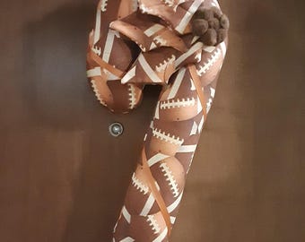May include: A candy cane shaped fabric decoration featuring a brown and tan football pattern with white laces. The top of the candy cane is adorned with a brown fabric bow and a small cluster of brown pom-poms, creating a sports-themed wall hanging.