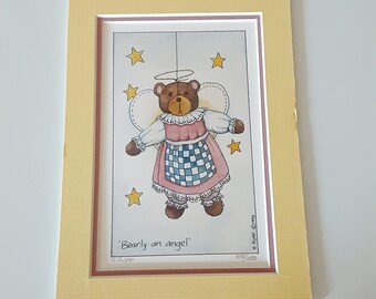 May include: A framed print featuring a brown teddy bear angel with a halo, wings, and a pink and blue checkered apron. The artwork is surrounded by yellow stars and framed in a yellow mat.