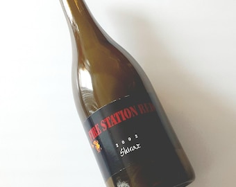 May include: A dark amber glass wine bottle with a black label featuring the red text FIRE STATION RED, the year 2002, and the word Shiraz in white script. A small graphic of a stylized figure is visible on the lower left side of the label.