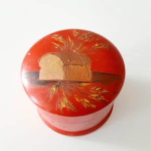 May include: A round, red container with a lid, featuring a hand-painted design of a loaf of bread and wheat stalks. The bread is painted in shades of brown and tan, with yellow and brown wheat stalks. The container is likely made of ceramic or a similar material.