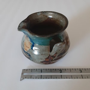 May include: A small ceramic creamer with a pour spout and a side handle, featuring a mottled glaze of blue, brown, and grey tones. A metal ruler placed in front shows the vessel has a width of approximately 2.5 inches at its widest point.