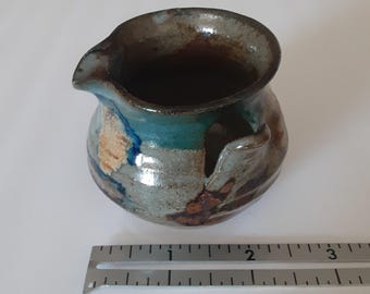 May include: A small ceramic creamer with a pour spout and a side handle, featuring a mottled glaze of blue, brown, and grey tones. A metal ruler placed in front shows the vessel has a width of approximately 2.5 inches at its widest point.