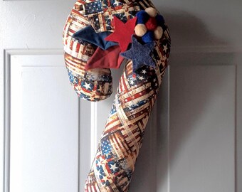 May include: A fabric candy cane decoration featuring a printed pattern of American flags with red, white, and blue stripes and stars. The top is adorned with layered felt stars in red and navy blue, along with a cluster of small, round felt balls.
