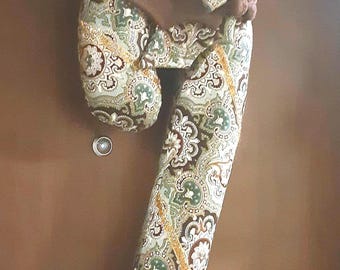 May include: A fabric candy cane decoration featuring a green, brown, and cream paisley pattern. The item is accented with a gold-colored ribbon trim and a small brown pom-pom at the top curve. A small silver-colored circular button is placed nearby.