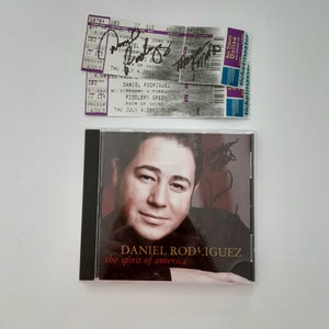 May include: A black CD case with a photo of Daniel Rodriguez on the front. The title of the CD is "The Spirit of America". There are two concert tickets for Daniel Rodriguez on the top of the CD case.