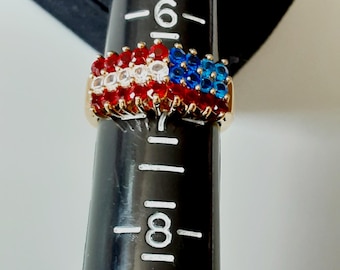 May include: A gold-toned ring featuring a design resembling the American flag. The ring's top is adorned with rows of red, white, and blue gemstones. The gemstones are arranged to mimic the stars and stripes of the flag. The ring is on a black ring sizer.