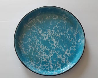 May include: A round enamel tray featuring a vibrant turquoise blue base color with a white marbled or swirled pattern throughout the surface. The tray has a thin, dark black rim that encircles the entire outer edge of the circular metal object.
