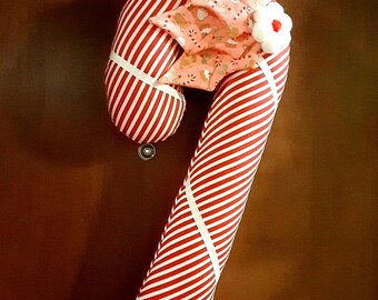 May include: A fabric candy cane decoration featuring a red and white diagonal striped pattern. The candy cane is accented with a white ribbon wrapped around it and a decorative fabric holly leaf topper with a small white flower and red center.