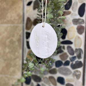 May include: A white, round, ceramic ornament with embossed botanical designs, hanging from a natural twine string. The ornament features a wheat stalk and other plant elements. Green eucalyptus leaves are in the background.