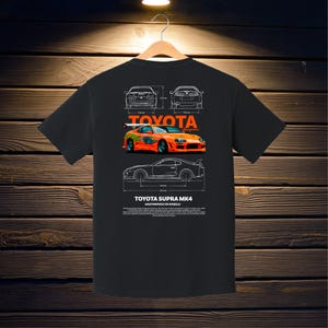May include: Black t-shirt featuring a graphic of a Toyota Supra MK4 car in orange and green, with white blueprint-style diagrams and the Toyota logo. The text "TOYOTA SUPRA MK4 MASTERPIECE ON WHEELS" is also printed on the shirt.