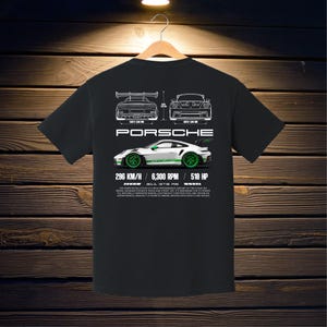May include: Black t-shirt featuring a white and green Porsche 911 GT3 RS graphic. The design includes car schematics, the Porsche logo, and performance specs: 296 KM/H, 6,300 RPM, and 518 HP. The shirt is hanging on a wooden hanger.
