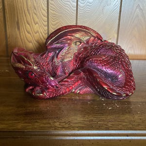 May include: A red dragon figurine with gold accents. The dragon is curled up in a sleeping position with its wings spread out.
