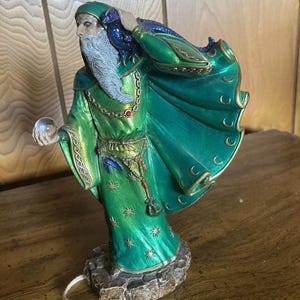 May include: A green and gold wizard figurine with a long white beard, holding a crystal ball in one hand and a raven on his shoulder. He is wearing a green robe with gold trim and a chain with a red jewel around his neck.