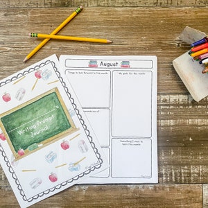 May include: A writing prompt journal and a blank monthly planner are displayed on a wooden surface, with two pencils and a container of crayons. The journal cover features a chalkboard design with apples, pencils, and erasers. The planner is labelled "August."