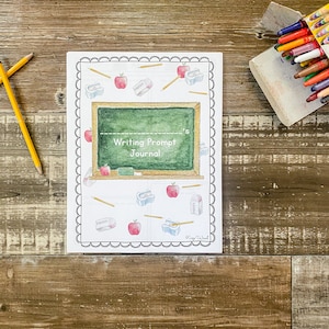 May include: A journal with a white cover and a black scalloped border. The cover features a green chalkboard design with the text "Writing Prompt Journal." It is decorated with watercolour illustrations of apples, pencils, erasers, and pencil sharpeners. Two pencils are on the left.
