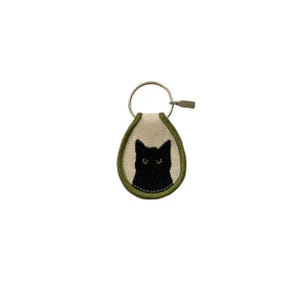May include: A keychain featuring an embroidered black cat design. The cat is set against a cream-colored background with a green border. The keychain includes a metal ring and clasp for attaching keys.