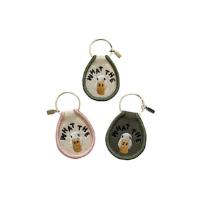 May include: Three embroidered keychains with a duck design and the text "WHAT THE". The keychains are in shades of green, pink, and beige, each with a gold-colored keyring and clasp. The duck has a yellow beak and is centered on the keychain.