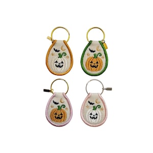 May include: Four teardrop-shaped embroidered keychains featuring jack-o'-lantern pumpkins, bats, stars, and crescent moons. The keychains have gold or silver metal rings and are bordered in orange, green, light purple, or light pink fabric trim.