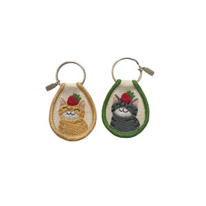 May include: Two embroidered keychains featuring cat faces wearing strawberry hats. One keychain has an orange cat with a gold border, and the other has a gray cat with a green border. Both have silver key rings.