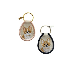 May include: Two embroidered corgi keychains. One keychain has a gold ring and a pink border, the other has a silver ring and a navy blue border. Both feature a corgi face.