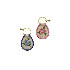 May include: Two embroidered keychains. One keychain features a blue floral design with a purple border, and the other has a pink floral design with a pink border. Both keychains have a gold-colored ring and clasp.