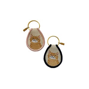 May include: Two embroidered keychains featuring a smiling orange tabby cat. One keychain has a pink border, the other a black border. Both have a gold-colored key ring and a small, gold-colored zipper pull.