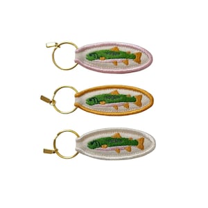 May include: Three embroidered keychains with a fish design. Each keychain features an oval shape with a gold-colored key ring. The fish is green with orange fins and is set against a white background with a pink, gold, or white border.