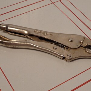 May include: A silver locking pliers tool with a textured handle and adjustable screw. The jaws are serrated and designed for gripping. The tool is made of metal and has a spring mechanism.