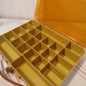 Puede incluir: Una caja de aparejos Plano de color amarillo mostaza abierta con una tapa naranja transparente. El interior está dividido en numerosos compartimentos de varios tamaños. Una correa de cuero marrón con un cierre de metal está sujeta al lateral.