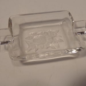 Op de afbeelding: Rechthoekige schaal van helder glas met een bloemenontwerp in het midden. De schaal heeft twee kleine handvatten aan weerszijden. Transparant glas, ideaal voor het presenteren van snoep of als decoratief element.
