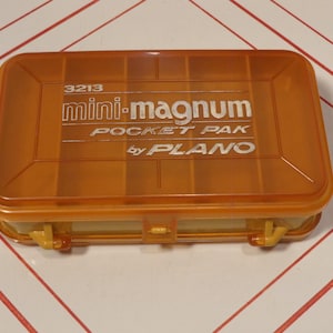 Puede incluir: Una caja de aparejos Plano Mini-Magnum Pocket Pak de plástico naranja. La caja tiene forma rectangular con tapa abatible y cierre amarillo. El texto "3213 mini-magnum POCKET PAK by PLANO" está impreso en la tapa.