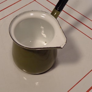 May include: Olive green enamelware saucepan with a white interior and spout. The handle is black and green. The pot is sitting on a white surface with red stripes.