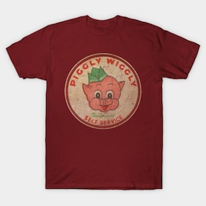 Peut inclure: T-shirt bordeaux avec un motif de style vintage. Le motif est un design circulaire avec les mots "PIGGLY WIGGLY" sur le bord et un cochon de dessin animé portant un chapeau vert au centre. Le texte "The Original Self Service" est en dessous.
