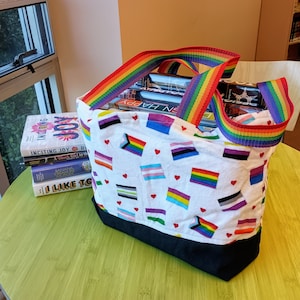 May include: A white tote bag with a rainbow strap and a black bottom. The bag is filled with books and has a pattern of different pride flags on the front. The bag is sitting on a green table.