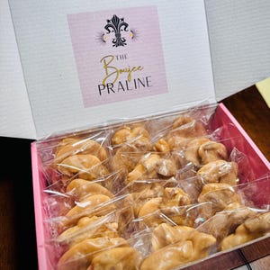 May include: A pink box filled with individually wrapped pralines. The pralines are a light brown color and appear to be made of a caramel or toffee-like substance. The box has a label that reads "The Boujee Praline".