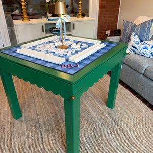 May include: A square green game table with a scalloped apron and straight legs. The tabletop features a blue and white plaid surface with white tile racks, a collection of letter tiles, and a small gold lamp with a light blue ribbon tied to it.