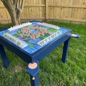 May include: A dark blue square game table with four legs, each featuring a circular cork inlay and a small gold-toned ball accent. The tabletop surface displays a colorful floral pattern and is set up with white tile racks and mahjong tiles.