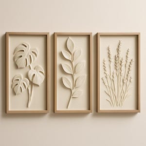 May include: Three framed wall art pieces with botanical designs. The art features a monstera plant, a leafy branch, and a cluster of wheat-like plants, all in cream. The frames are light brown.